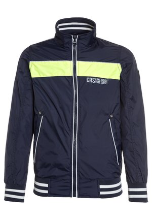 Chaqueta azul marino con una franja horizontal amarilla, detalles en blanco, cierre de cremallera y puños a rayas. Hecha de un material ligero y suave.