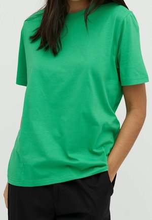 Grünes Baumwoll-T-Shirt mit kurzen Ärmeln, lässiger Passform, Rundhalsausschnitt, glatter Struktur und abgesteppten Säumen. Kombiniert mit schwarzen Hosen, Hände in den Taschen.