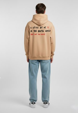 Felpa beige con cappuccio e testo nero e rosso sul retro. Abbinata a pantaloni azzurri chiari e sneakers bianche e nere. Texture liscia.