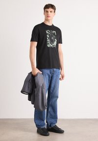 Zwarte katoen t-shirt met een groene grafische ontwerp op de voorkant, gecombineerd met blauwe jeans en een grijze, gestructureerde jas die in één hand wordt vastgehouden.