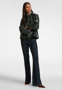 Giacca in pelle verde con bottoni a scatto, vestibilità rilassata e camicia blu sotto. Abbinata a jeans svasati scuri e tacchi marroni.