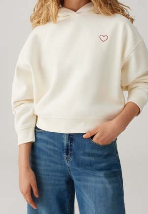 Personne portant un sweat à capuche crème avec un petit cœur rouge sur la poitrine et un jean bleu, une main dans la poche.