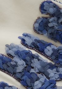 Tekstiludsmykning med lagde blå stofblade med en glat overflade, kantet med små lilla perler på en cremefarvet baggrund.