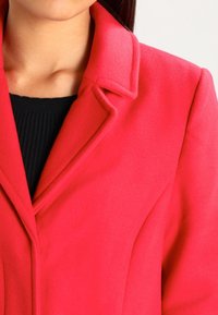 Abrigo de lana rojo brillante con cuello con muescas, ajuste entallado y superficie texturizada, llevado sobre una camiseta negra de canalé.
