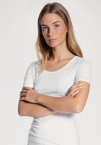 White fitted t-shirt with short sleeves, round neckline, smooth texture, and stretchy fabric. Worn by a model with arms crossed.