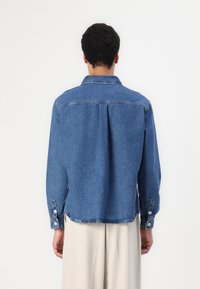 Giacca di jeans con vestibilità morbida, colore azzurro chiaro, colletto classico e polsini con bottoni; l'orlo grezzo e il dettaglio del giogo posteriore arricchiscono il design.
