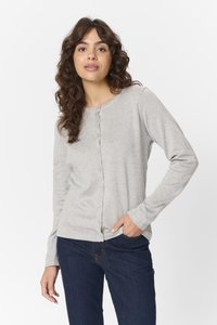 Žena s hnědými kudrnatými vlasy, oblečená v světle šedém knoflíkovém kardiganu a tmavě modrých džínách, stojící s jednou rukou v kapse na bílém pozadí.