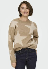 Beige trui met grote crèmekleurige sterren, ronde halslijn en geribde manchetten. Gepaard met donkerblauwe jeans met een gestructureerd breiwerk ontwerp.