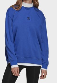 Felpa di cotone blu con maniche lunghe, polsini a costine e scollo rotondo. Presenta un piccolo logo Adidas nero sul petto.