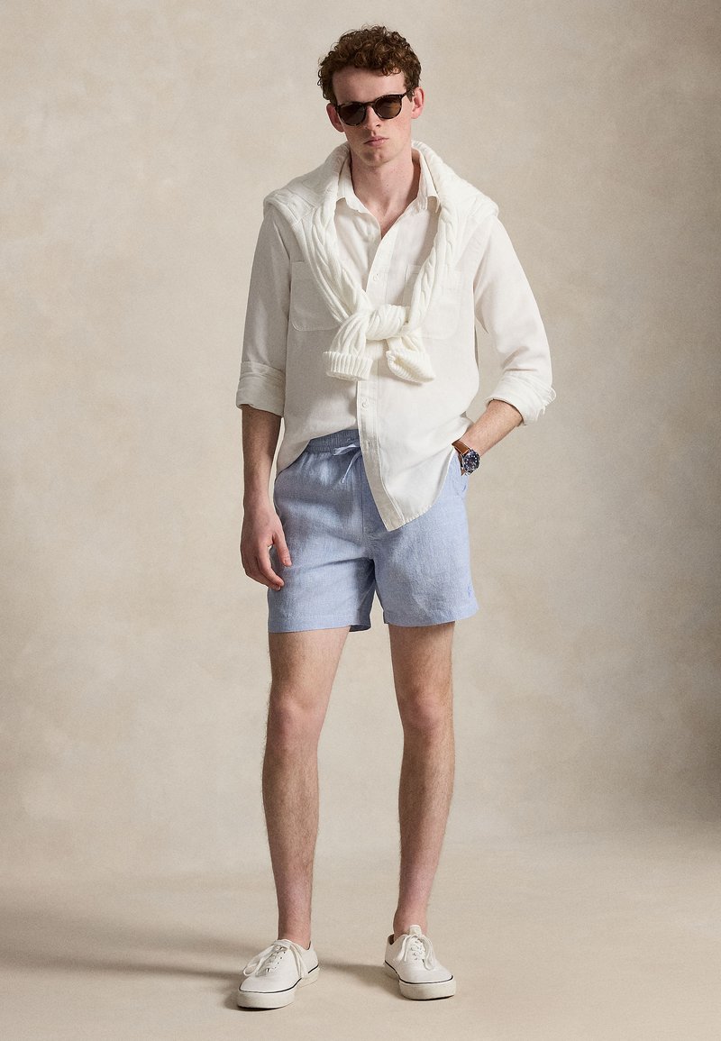 Jeune homme portant des lunettes de soleil, une chemise blanche boutonnée avec un pull blanc noué aux épaules, un short bleu clair, des baskets blanches et une montre.