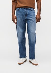 Uomo che indossa jeans azzurri a gamba dritta, maglione marrone a maniche lunghe e sneakers bianche con suole marroni, in piedi davanti a uno sfondo semplice.