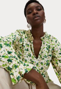 Blusa a trama floreale verde e bianca con scollatura a V, colletto arricciato e maniche a palloncino. Tessuto leggero con una sottile texture.