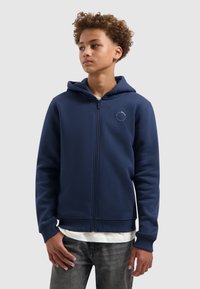 Maritime ritsjack in zachte stof met een voorzak, ribbelmanchetten en een ronde logo op de borst. Gelaagd over een wit shirt.