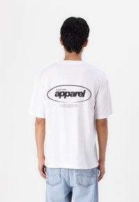 Camiseta de algodón blanca de manga corta, con un gran logo negro en la parte de atrás y un texto debajo. Corte recto, diseño minimalista.