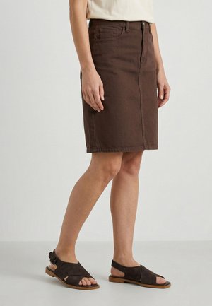 Persoon draagt een knielange bruine denim rok en donkerbruine open-toe sandalen met gespen op een neutrale achtergrond.