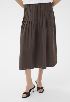 Jupe midi plissée marron avec des détails œillets le long de l'ourlet, portée avec des sandales à talons hauts noires à bout ouvert sur fond blanc.