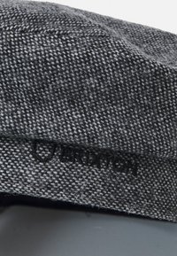 Casquette grise à motif en tissu texturé, comportant un petit logo imprimé sur le côté. Détails de couture visibles sur l'ensemble du matériau.