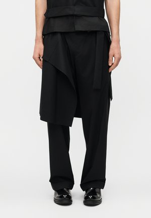 Άτομο που φορά μαύρο παντελόνι σε στρώσεις με ασύμμετρο draped πάνελ πάνω από παντελόνια με ίσια πόδια και μαύρα γυαλιστερά παπούτσια.