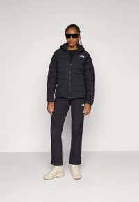 Svart pufferjacka med dragkedja fram och huva, bärs över en svart topp och byxor. Beige sneakers kompletterar outfiten.