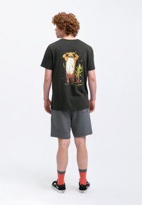 Zwarte katoenen t-shirt met een kleurrijke berenafbeelding op de rug, gecombineerd met grijze shorts, rode sokken en zwarte sneakers.