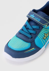 Modrá sportovní bota vyrobená z prodyšných a syntetických materiálů, s texturovanou špičkou, polstrovaným jazykem a nastavitelným popruhem s logem.