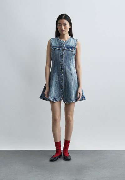 Νεαρή γυναίκα που στέκεται φορώντας αμάνικο τζιν φόρεμα με τρουκς, κόκκινες κάλτσες και μαύρα flats, μπροστά σε απλό γκρι φόντο.