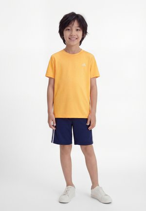 Fant, ki stoji in se smeji, oblečen v oranžno majico Adidas, temno modre kratke hlače z belimi črtami in bele superge, na belem ozadju.