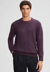 Maglione viola con texture, collo a giro e orlo a coste, abbinato a pantaloni neri. Il tessuto sembra morbido e leggermente peloso.