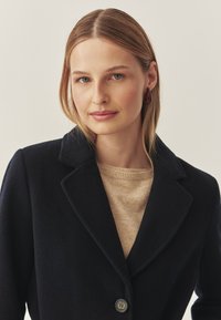 Marineblauer Wollmantel mit Reverskragen und einfacher Knopfschluss, kombiniert mit einem hellbeigen Pullover. Glatte Textur mit klassischem Design.