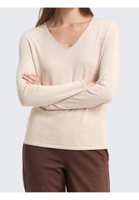 Haut beige à manches longues avec col en V, fabriqué en tissu doux, présentant une coupe décontractée et une texture subtile. Associé à un pantalon marron.