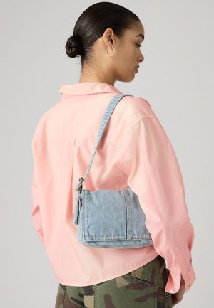 Donna con capelli scuri raccolti in uno chignon, che indossa una maglietta rosa e pantaloni mimetici, con una borsa a tracolla in denim azzurro chiaro sulla schiena.