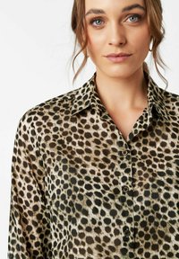 Woman with light skin and blue eyes wearing a long-sleeve leopard print button-up shirt against a plain white background.