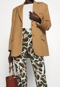 Veste marron avec des revers crantés, associée à un pantalon à motifs avec des tons verts et beiges. Le mannequin tient un sac à bandoulière marron.