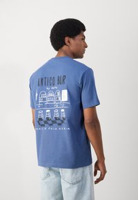Blauwe katoenen T-shirt met een grafische print van een bar scène op de achterkant, met witte tekst en illustraties van barkrukken. Normale pasvorm.