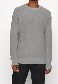 Maglione grigio lavorato a maglia con scollo rotondo, polsini e orlo a coste. Tessuto testurizzato con una vestibilità morbida, mostrato abbinato a pantaloni neri.