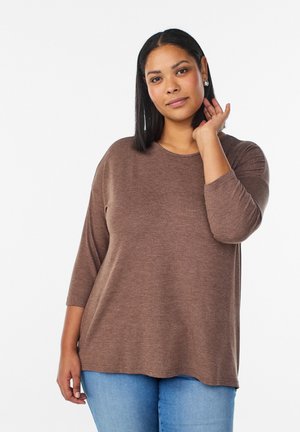 Kobieta o średnim odcieniu skóry i prostych czarnych włosach, ubrana w brązową bluzkę z długim rękawem i jasnoniebieskie dżinsy, dotykająca swoich włosów.