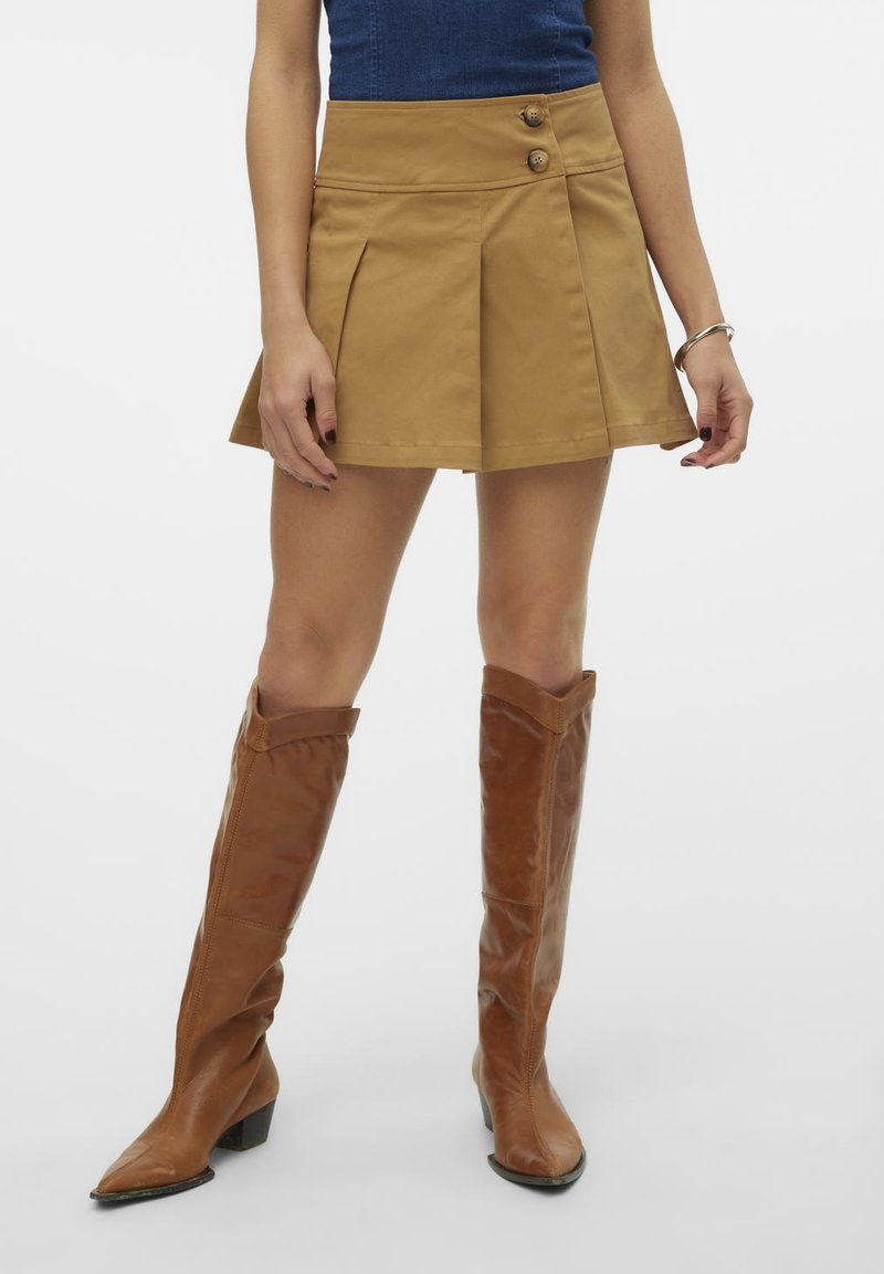 Beige plissert skjørt med to knapp detaljer, laget av et teksturert stoff. Kombinert med høye brune lærstøvler.