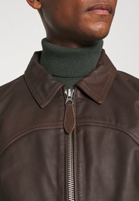 Man som bär en mörkbrun läderjacka med en silverdragkedja över en grön ribbad polotröja.