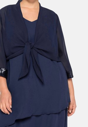 Femme portant une robe bleu marine avec un cardigan transparent assorti noué à la taille et des poignets à sequins aux manches trois quarts.