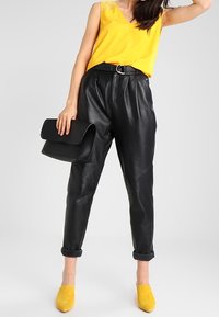 Pantalons en cuir noir avec des plis, un top sans manches jaune, des chaussures en suède jaune et un sac à main noir avec une finition texturée.
