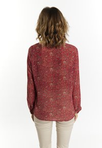 Blusa rossa con motivo floreale, maniche lunghe e vestibilità rilassata, realizzata in tessuto testurizzato e con dettagli sottili in sfumature di oro e blu.