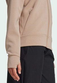 Detailný záber na osobu v béžovej mikine na zips s otvormi na palce a čiernymi nohavicami, zobrazený detail zipsu bočnej vrecka.