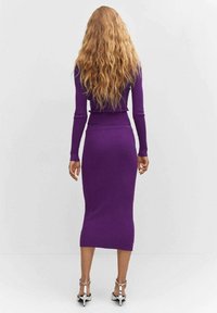 Femme aux longs cheveux blonds ondulés se tient face à un mur, portant une robe moulante à manches longues en purple côtelé et des sandales à talons hauts argentées.