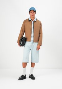 Young man wearing tan jacket, striped shirt, light denim shorts, white socks, black loafers, blue cap, holding black clutch against white background.