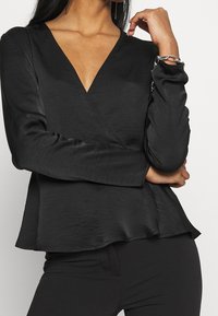 Černá blůza s hlubokým výstřihem do V, dlouhými rukávy a rozšířeným lemem. Vyrobena z hladké, strukturované látky. Obsahuje stříbrný řetízek jako akcent na zápěstí.