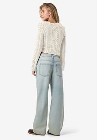 Cremefärgad stickad kort svetttröja med diamantmönster och långa ärmar, tillsammans med ljusblå vida jeans. Bakifrån visas fickdetaljer.