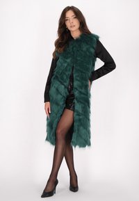 Teal-farbene Kunstpelzweste mit langer, struppiger Textur, offener Vorderseite, getragen über einer schwarzen Bluse und glänzenden Shorts, kombiniert mit schwarzen High Heels.