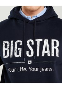 Vīrietis tērpies tumši zilā kapuci džemperī uz gaiši zila apkaklīša krekla, uz kapuces ir balts uzraksts "BIG STAR Your Life. Your Jeans."