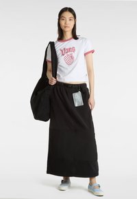 Hvid t-shirt med lyserød kant og grafisk design, parret med en lang sort nederdel og blå sko, bærende en sort taske med sidelomme.
