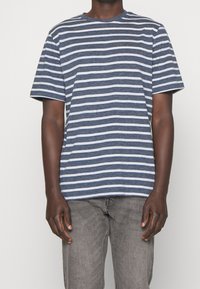 Modrá a bílá pruhovaná tričko s krátkým rukávem, kulatý výstřih, vyrobené z měkkého materiálu. V kombinaci se šedými džíny. Viditelné švy.